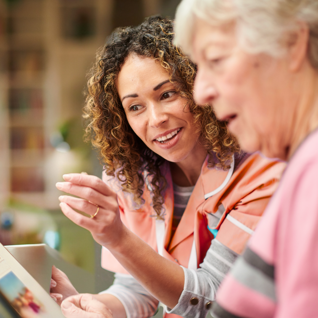 A woman with curly hair showing an elderly lady how to use the new client management software in a home
