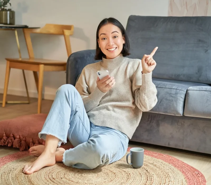 A woman sitting on the floor in a lounge room leaning against a grey couch pointing to the sky.